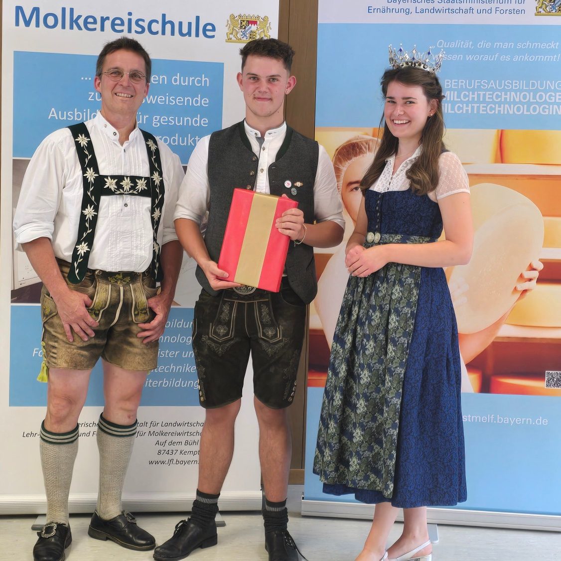 Drei Personen in traditioneller Kleidung stehen vor einem Banner der Molkereischule.